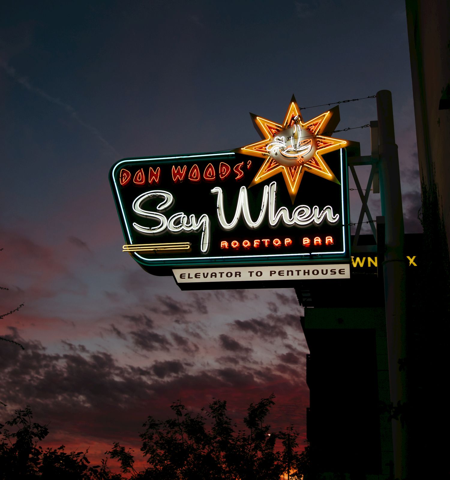 A neon sign for "Don Wood's Say When Rooftop Bar" with a star design is illuminated against a sunset sky.