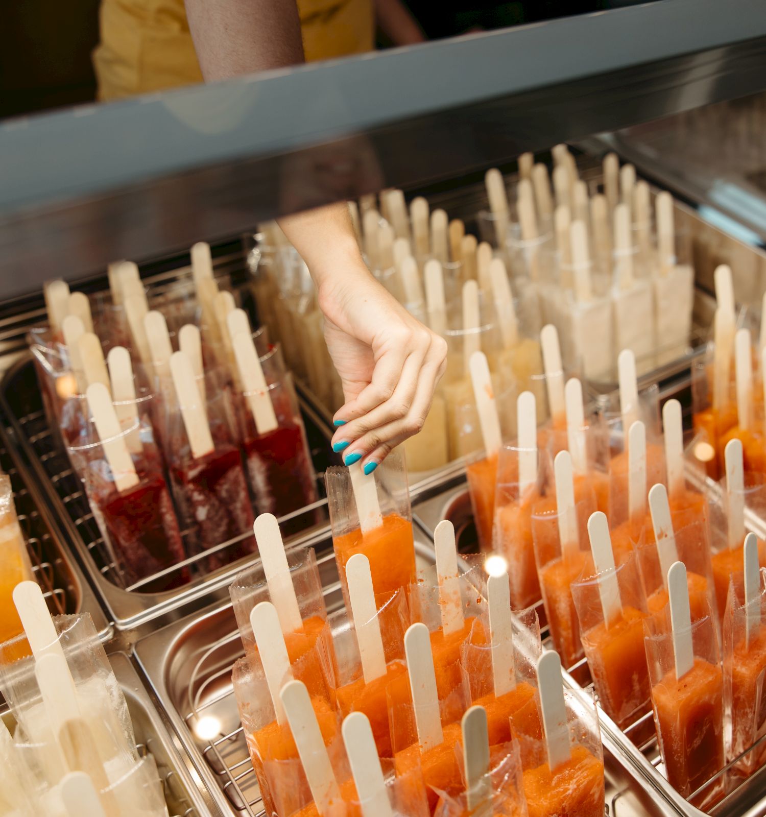 A person is choosing a popsicle from a display filled with various colorful popsicles in a shop.