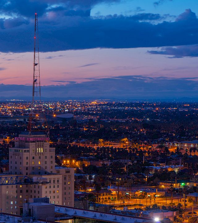A dusk city skyline with lights, a tall lattice broadcast tower, and stormy clouds over the horizon.