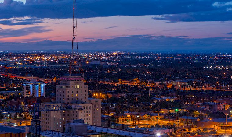 A dusk city skyline with lights, a tall lattice broadcast tower, and stormy clouds over the horizon.