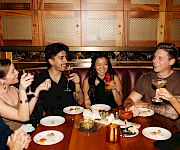 A group of friends in a cozy restaurant booth cheers with beer and laughs over plates at the table.