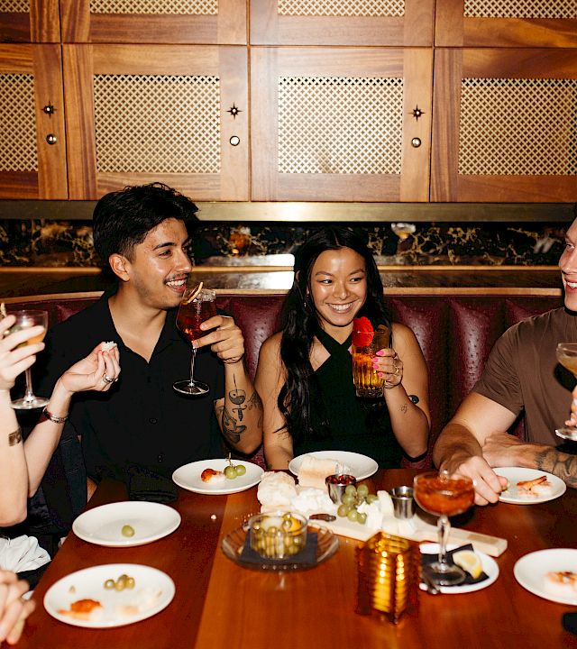 A group of friends in a cozy restaurant booth cheers with beer and laughs over plates at the table.