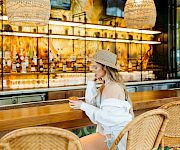 A woman in a straw hat relaxes at a wicker cafe table, sipping a drink under woven pendant lights.