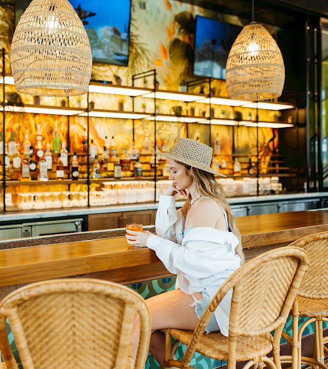 A woman in a straw hat relaxes at a wicker cafe table, sipping a drink under woven pendant lights.