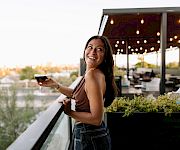 A woman smiles on a patio holding a glass of wine as warm lights glow behind her.