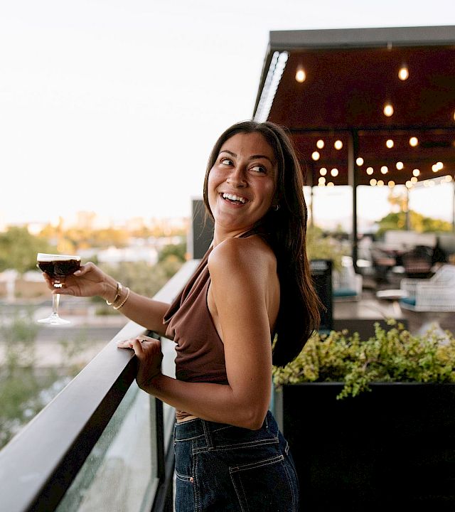 A woman smiles on a patio holding a glass of wine as warm lights glow behind her.