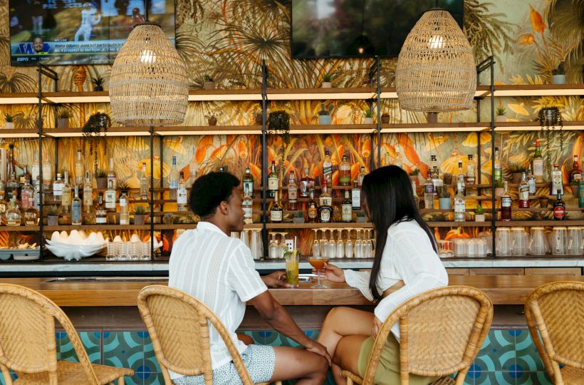 A couple sits at a tropical bar with cocktails, lots of liquor bottles, and rattan lamps under leafy wall art.